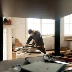 Pracownik w ochronnych okularach i słuchawkach szlifuje metalowe elementy. W tle widać okna i materiały budowlane, co sugeruje modernizację starego domu.