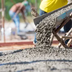 Beton wylewany z rynny na placu budowy. W tle pracownicy. Obrazek pomoże zrozumieć, jak obliczyć ile kubików betonu potrzeba.