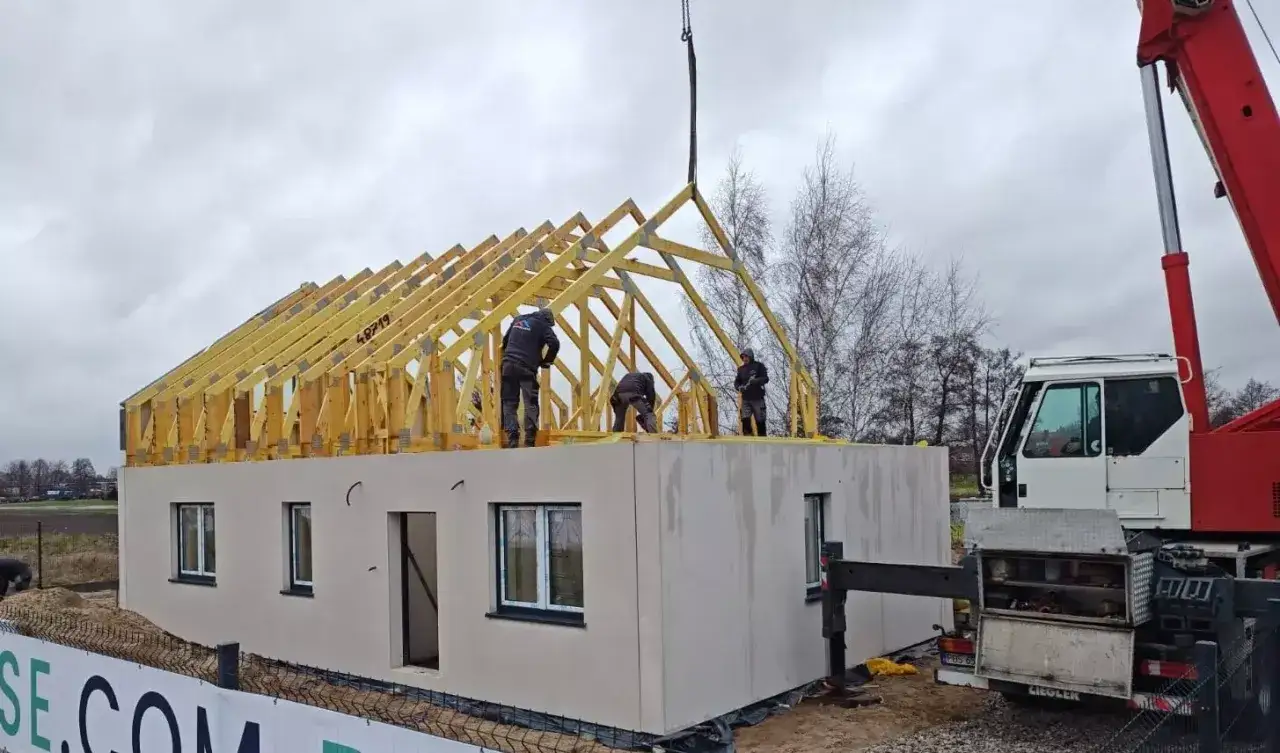 Montaż prefabrykowanych wiązarów dachowych na budowie domu. Pracownicy i dźwig na placu budowy.